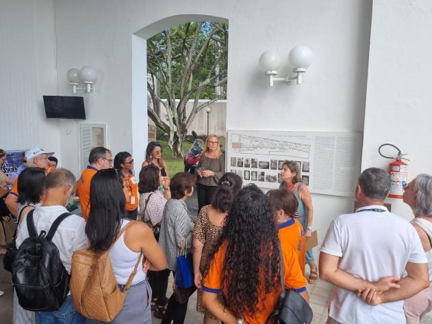 Grupo de visitantes reunidos em área expositiva do Cemitério Britânico, ouvindo explicações de duas diretoras diante de um painel informativo.