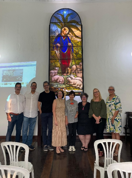 Grupo de pessoas posando diante de um grande vitral colorido em uma sala interna, após atividade no Museu e no Cemitério Britânico.
