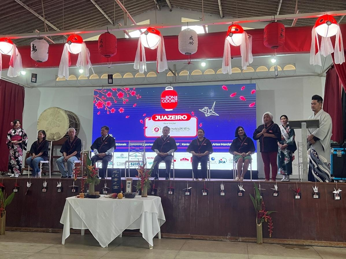 Abertura do I Bon Odori Itinerante da Bahia em Juazeiro, com organizadores no palco diante de um painel iluminado e decoração japonesa.