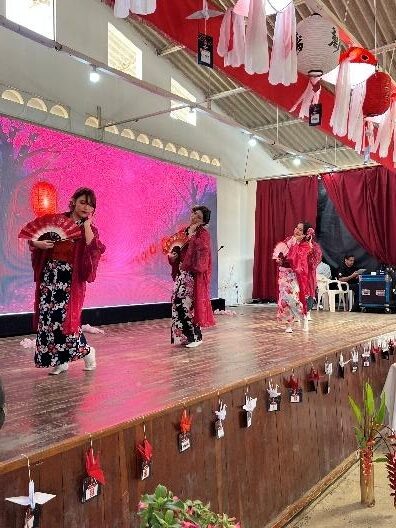 Apresentação do grupo de dança Hanabi no palco, com três integrantes usando quimonos floridos e segurando leques diante de um painel iluminado em tons de rosa.