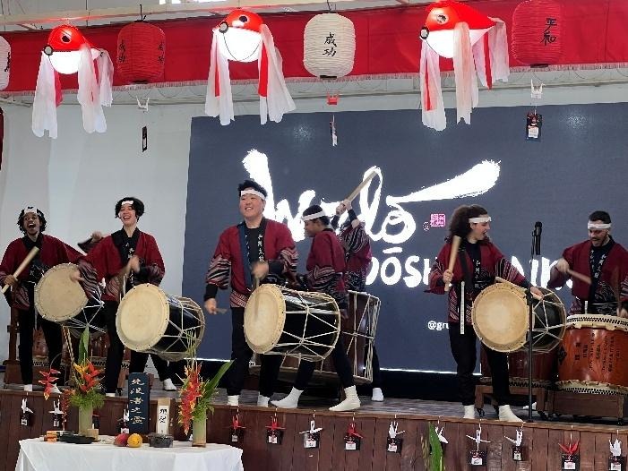 Grupo de taikô Wado se apresenta no palco, com percussionistas usando trajes tradicionais e tocando tambores japoneses diante de um painel iluminado.