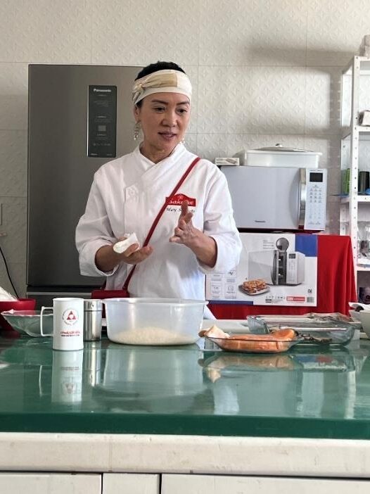 Chef Mary Sugimoto conduz oficina de culinária, posicionada atrás de uma bancada com ingredientes e utensílios.