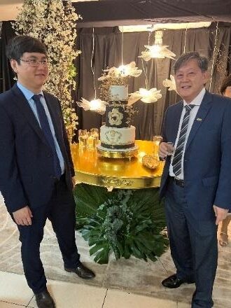 Dois homens em trajes formais posam diante de mesa decorada com bolo comemorativo e arranjos florais, durante celebração institucional em ambiente interno.