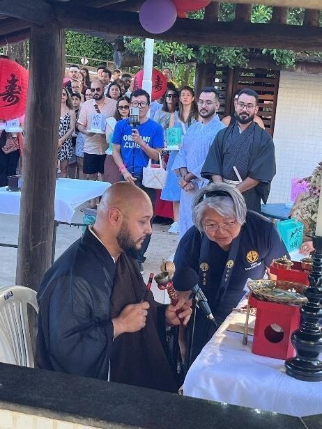 Monge conduz ritual ecumênico japonês do Tōrō Nagashi diante de altar cerimonial, enquanto participantes acompanham a celebração em espaço ao ar livre.