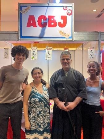 Quatro participantes posam em frente ao estande da ACBJ-BA durante evento cultural japonês, reunindo músico, artista literária, representante institucional e violinista.