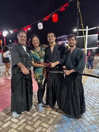 Quatro pessoas posam juntas durante o Bon Odori, sendo três praticantes de artes marciais japonesas com trajes tradicionais e uma representante institucional, em ambiente noturno decorado com lanternas.