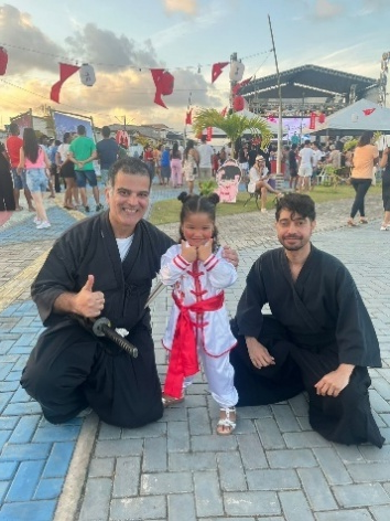 Homem e jovem praticante de artes marciais japonesas ajoelhados ao lado de uma criança vestida com traje tradicional, durante o Bon Odori, em espaço aberto com decoração festiva.
