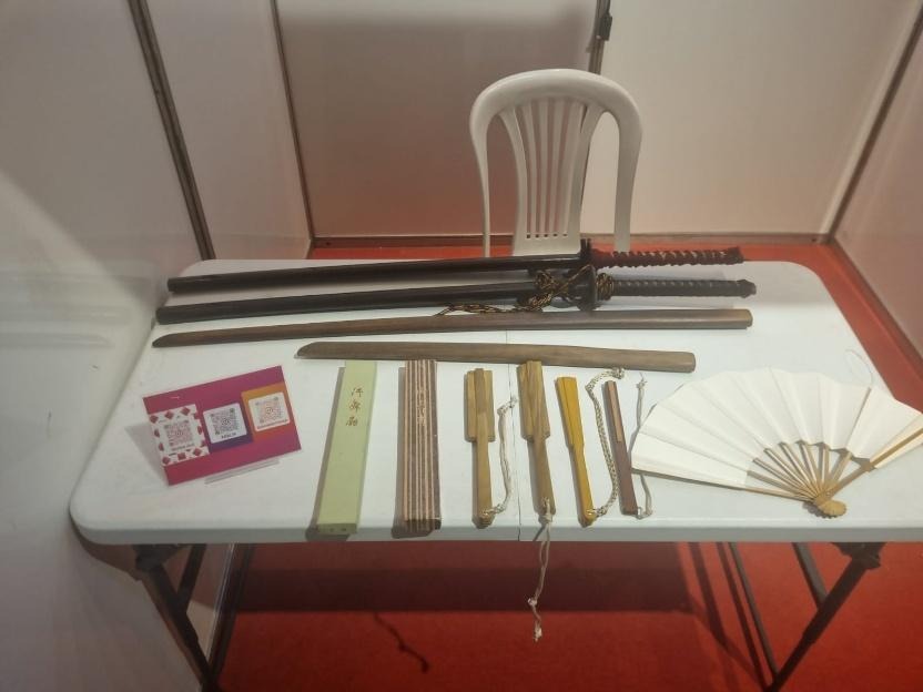 Mesa expositiva com espadas japonesas, bainhas, peças de madeira, ferramentas artesanais e leque tradicional, apresentados em estande cultural.