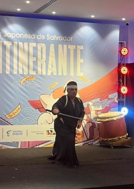 Praticante de arte marcial japonesa realiza apresentação com espada durante demonstração do Shinto Ryu Iai Battojutsu em palco de evento cultural.