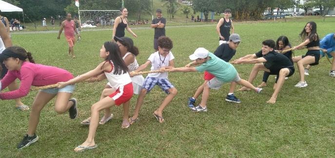 Crianças, adolescentes e adultos participam de provas tradicionais do Undokai, incluindo o Tsunahiki (cabo-de-guerra), em área aberta e gramada, promovendo integração e espírito de equipe.