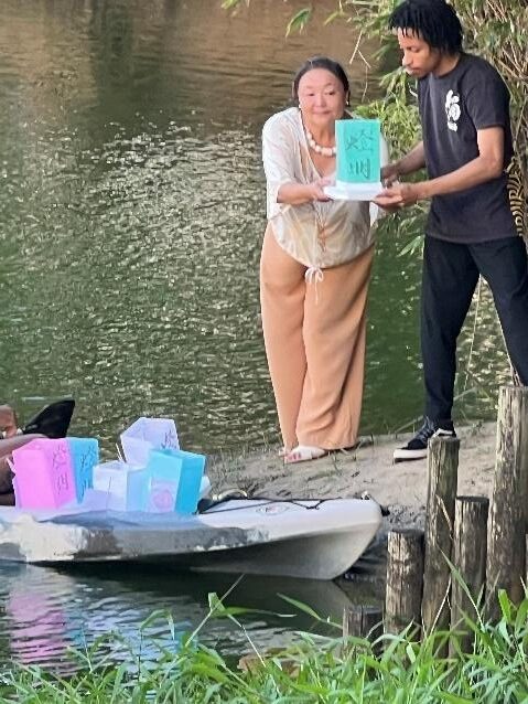 Mulher e jovem seguram lanternas de papel com mensagens antes de colocá-las na água durante o Tōrō Nagashi, em ambiente natural junto à lagoa.