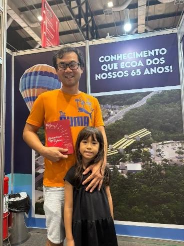Fotografia de Marcos Shimizu, homem de óculos e camiseta laranja da Puma, sorrindo ao lado de sua filha Luísa, uma menina de cabelos longos e vestido preto. Ele segura o livro de capa vermelha "Singulares Relações Nipo-Baianas". Eles estão em pé dentro do estande, em frente a um painel com a inscrição "Conhecimento que ecoa dos nossos 65 anos!".
