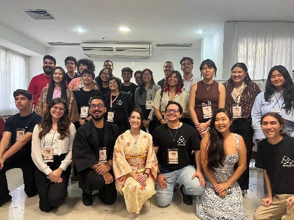 Fotografia de um grupo grande e diversificado de jovens posando sorridentes para a câmera em uma sala. Todos usam crachás do evento no pescoço. No centro da primeira fileira, destacando-se, há uma jovem vestindo um quimono tradicional de cor clara com estampas florais.