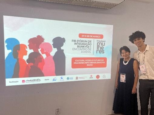 Fotografia de uma mulher e um jovem rapaz posando sorridentes, lado a lado. Eles usam crachás do evento. Ao lado deles, ocupando a maior parte do lado esquerdo da imagem, há uma tela de projeção iluminada com o painel do evento Fórum de Integração Bunkyo (FIB).