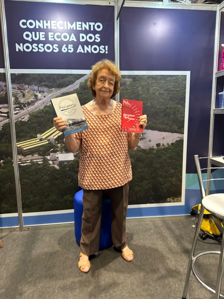 Fotografia de corpo inteiro da Dra. Ogvalda Devay em pé no estande da Bienal. Ela sorri para a câmera enquanto segura o livro "Periperi" (capa branca) na mão direita e o livro "Singulares Relações Nipo-Baianas" (capa vermelha) na mão esquerda. Ao fundo, há um painel fotográfico com uma vista aérea e a inscrição institucional da universidade.