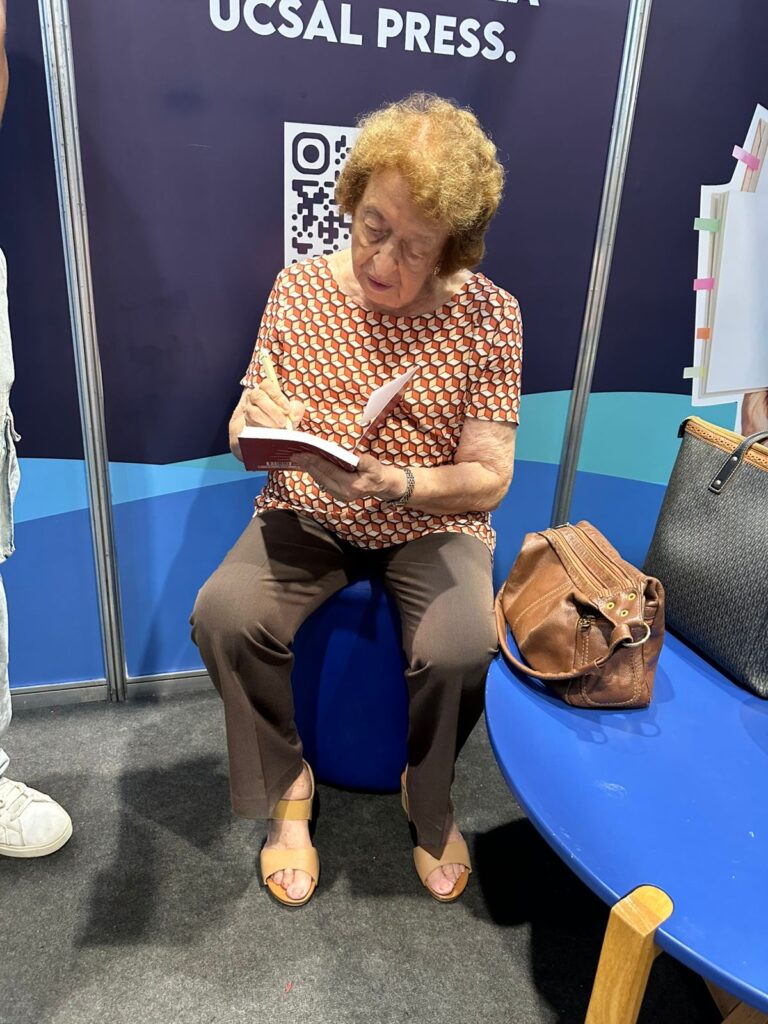 Fotografia colorida mostrando em plano médio a Dra. Ogvalda Devay sentada em um banco azul no estande da UCSal Press. Ela está concentrada, segurando uma caneta e assinando a página de rosto de um livro aberto. Ela veste uma blusa com padronagem geométrica e calça comprida marrom.